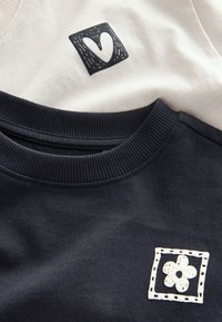 Két összehajtogatott pulóver, az egyik krémszínű fekete szívfolttal, a másik fekete fehér virágfolttal, mindkettő hímzett mintákkal a nyakrésznél.