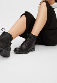 Schwarze Leder-Ankle-Boots mit einem Schnürverschluss, gepolstertem Kragen und einem Reißverschluss auf der Rückseite. Die Sohle ist robust und hat eine strukturierte Oberfläche.