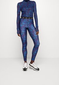 En person står klädd i en blå och lila mönstrad polotröja och matchande leggings, kombinerat med svarta och vita träningsskor.