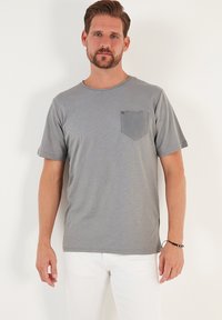 Siva kratka majica s kratkimi rokavi in okroglim izrezom ter potiskanim žepom na levi strani. Narejena iz mehkega materiala, sproščen kroj.