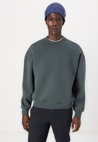 Mörkgrön sweatshirt med rundad halsringning, ribbade ärmslut och nederkant. Modellen bär en blå mönstrad stickad mössa. Enfärgad och avslappnad design.