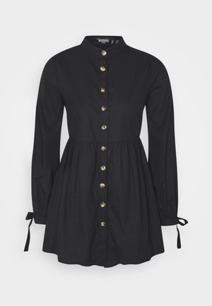 Robe noire à boutons, manches longues avec des détails à nouer, taille cintrée, en tissu doux. Présente des boutons dorés sur le devant.