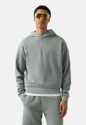 Jonge man met een saliegroene hoodie en bijpassende joggingbroek, met oversized gele sportzonnebril, staand met handen in de zakken.