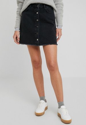 Vrouw draagt een zwarte denim rok met knopen aan de voorkant, een grijze trui met gestreepte manchetten, grijze sokken en witte plateau sneakers, staand tegen een effen achtergrond.