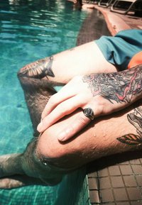 Hopeinen sormus, jossa on pyöreä musta yksityiskohta, lepää kädessä tatuoidun jalan päällä. Jalka on osittain upotettuna sinivalkoisilla laatoilla päällystettyyn uima-altaaseen.