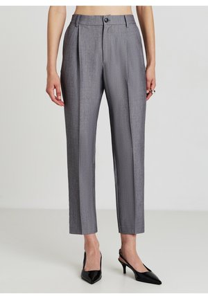 Kvinde iført grå højtaljede skræddersyede bukser med folder foran og sorte spidse slingback hæle, stående mod en ensfarvet lys baggrund.