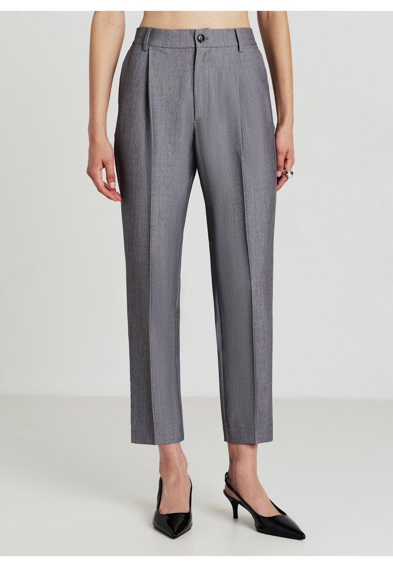 Femme portant un pantalon gris taille haute sur mesure avec plis à l'avant et des escarpins slingback noirs à bout pointu, debout devant un fond clair uni.