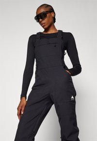 Black snow overalls with adjustable straps, front pocket, side zipper, and logo accent. Model wears a long-sleeve top and sunglasses.