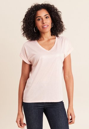 Woman with curly hair wearing hoop earrings, a light pink V-neck T-shirt, and dark jeans, standing against a beige background.