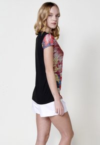 Top de manga corta con un frente colorido y estampado en tonos rojos, azules y dorados, con la parte trasera negra y un cuello redondo. Combinado con pantalones cortos blancos.