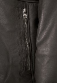 Nahaufnahme einer schwarzen Lederjacke mit einer Reißverschlusstasche am Arm, strukturiertem Material und vertikalen Nahtdetails entlang des Reißverschlusses.