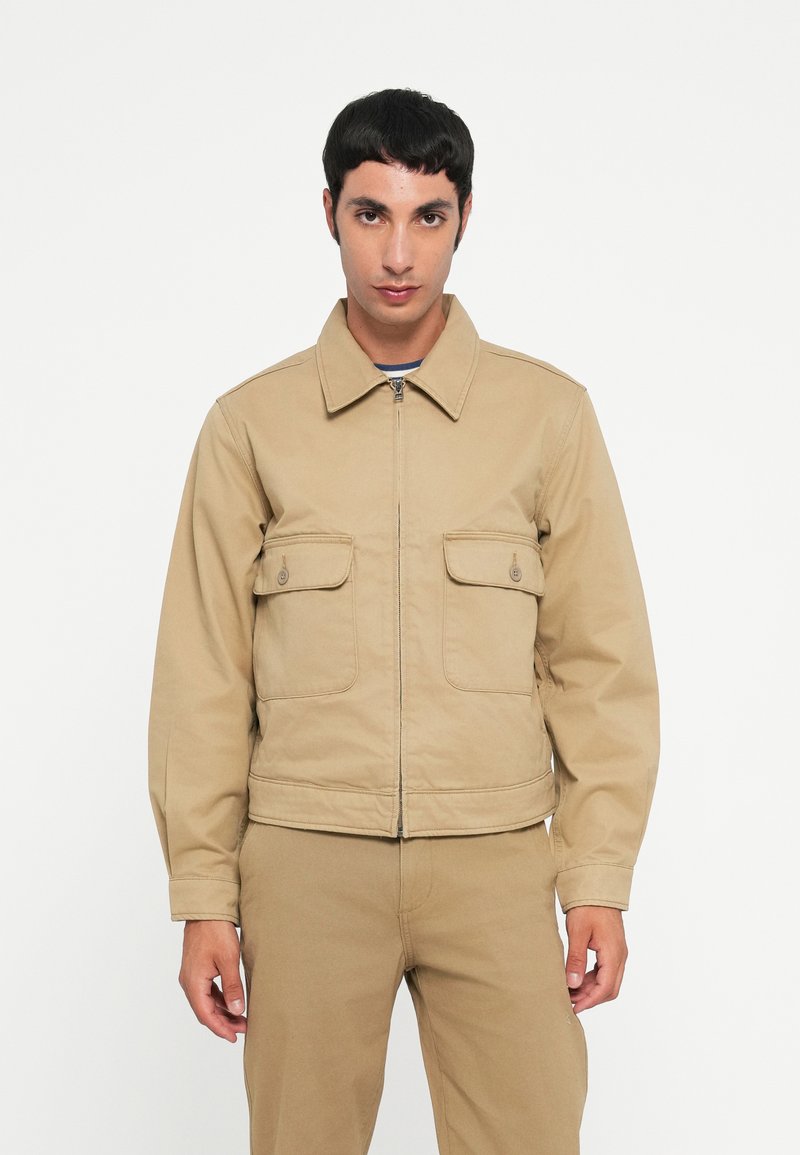 Beige cotton jacket with a zip front, pointed collar, and two chest pockets. Features long sleeves and a cropped design.