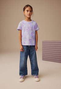 Cămașă mov tie-dye cu mâneci scurte și modele florale, asortată cu blugi evazați din denim și adidași cu modele. Textură netedă a țesăturii.