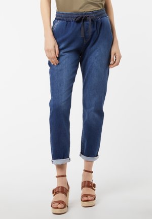 Kvinde iført opsmøgede blå denim joggebukser med elastisk talje og brune kilehælssandaler med ankelstropper.