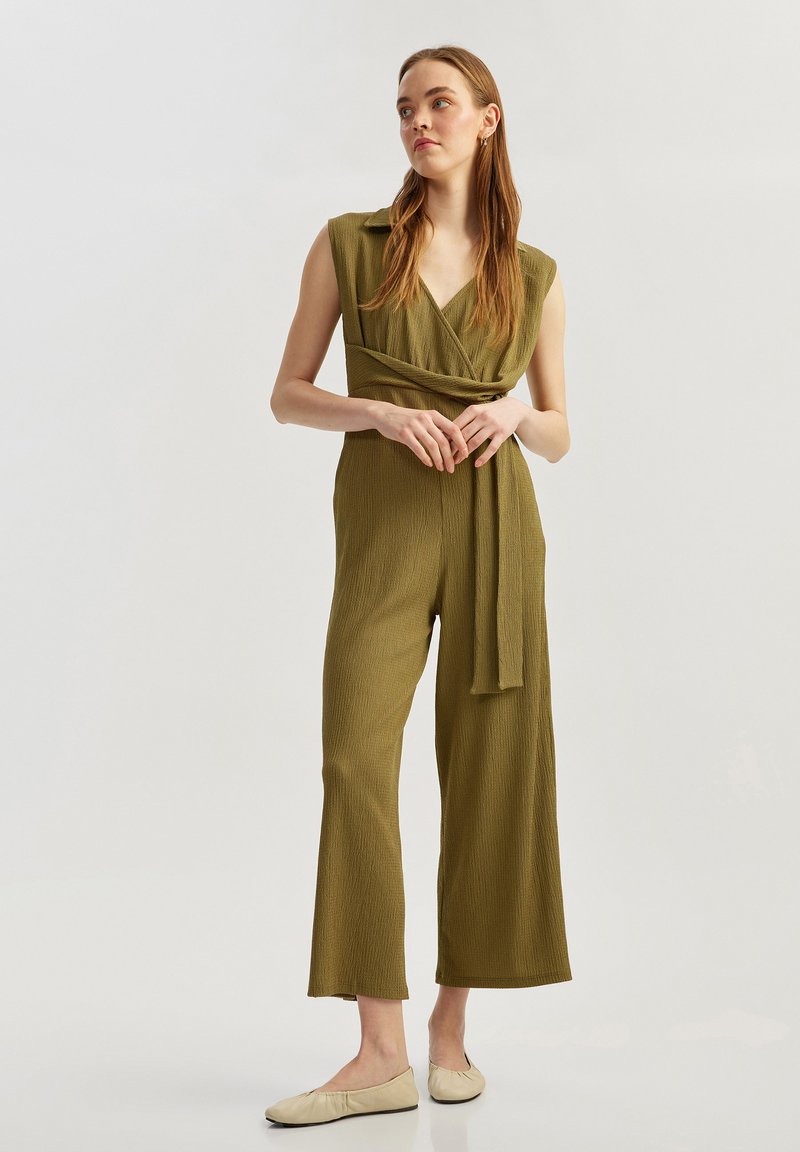 Olivgrön, ärmlös jumpsuit med V-ringning och midjebälte. Texturerad tyg, bred ben-design och släta beige ballerinaskor.