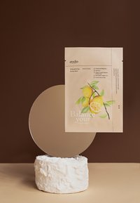 En förpackning med kol- och yuja-bubbelmask mot en brun bakgrund, med ljusa citronillustrationer, placerad på en strukturerad vit stensockel och spegel.
