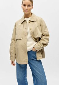 Femme portant une veste beige boutonnée, un pull en maille crème et un jean bleu, debout avec une main dans la poche devant un fond uni.
