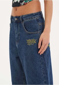 Dark blue denim jeans with a relaxed fit. Features yellow embroidered branding on the front pocket and classic five-pocket design.