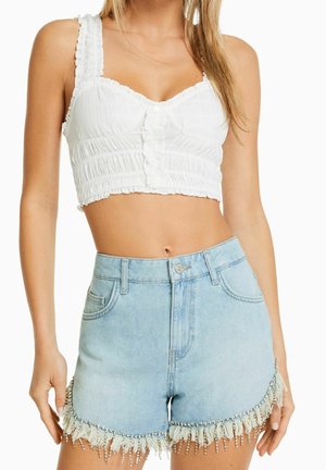 Frau trägt ein weißes, gerafftes Crop-Top mit Knöpfen und hellblaue Jeansshorts, die am Saum mit Fransen und Perlen verziert sind