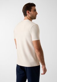 Jasnobe�żowa koszula z krótkim rękawem o prążkowanej fakturze i klasycznym okrągłym dekolcie, zestawiona z ciemnymi spodniami. Styl minimalistyczny.