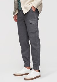 Homme portant un pantalon cargo jogger gris avec des poignets élastiques et des baskets blanches, debout avec une main dans la poche devant un fond uni.