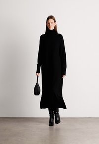 Μαύρο πλεκτό φόρεμα με μακριά μανίκια και ψηλό κόψιμο, με χαλαρή εφαρμογή, πλαϊνές σχισμές, το οποίο συνδυάζεται με μαύρες ankle μπούκες και μικρή τσάντα.
