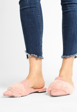 Fødder iført pink ruskind slip-on sko med blød pelskant, kombineret med blå cropped jeans med frynset kant på hvid baggrund.