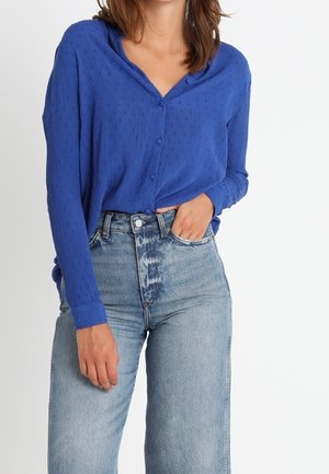 Kvinde iført en tekstureret blå knapbluse, der er stoppet ind i højttaljede lyseblå denimjeans, med den ene hånd i lommen.