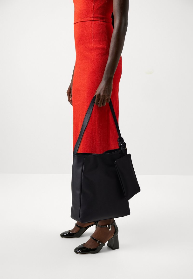 Personne portant une robe rouge, tenant un grand sac à main noir et portant des talons en cuir verni noirs avec des brides à la cheville.