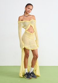 Rochie galbenă cu umerii descoperiți, marginile cu volane, material transparent, detaliu decupat la talie, mâneci lungi și fustă în straturi. Asortată cu adidași.