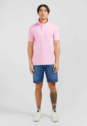 Polo shirt rose clair avec un col, associé à un short en denim et des baskets blanches. L'ensemble a des manches courtes et une coupe ajustée.