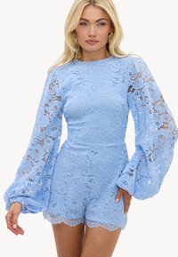 Jeune femme aux cheveux blonds portant une combinaison short en dentelle bleu clair avec des manches longues bouffantes et un ourlet festonné, se tenant devant un fond blanc.