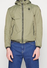 Chaqueta ligera de color verde oliva con capucha, frente con cremallera, bolsillos laterales, puños elásticos y detalles de costura en contraste.