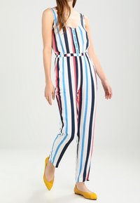 Randig jumpsuit med vertikala ränder i blått, rött och marinblått, med knäppning fram och åtsittande midja. Matchad med gula lägenheter.