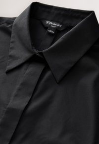 Chemise noire à col avec patte de boutonnage cachée, étiquetée "The Essential Shirt NEXT", fabriquée au Bangladesh.