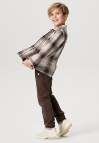 Geruite flanellen shirt in beige en bruin, grijze joggingbroek en lichtgekleurde sneakers. Kind staat zijwaarts, glimlachend, met de handen die het shirt vasthouden.