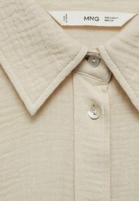 Chemise en tissu beige texturé avec un col pointu et deux boutons blancs visibles sur la patte de boutonnage avant.