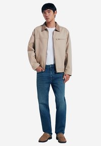 Hellbeige Jacke mit Reißverschlusstasche, getragen über einem weißen T-Shirt, kombiniert mit blauen Jeans und braunen Schuhen. Einfaches, strukturiertes Design.