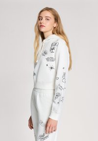 Felpa bianca corta con grafiche nere, tra cui un coniglio, stelle e testo. Abbinata a pantaloni della tuta bianchi coordinati che presentano grafiche simili.