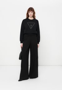 Mujer vistiendo sudadera negra de Guess, pantalones anchos negros con bolsillos, sosteniendo un bolso negro, de pie contra un fondo blanco.
