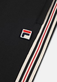 Zwarte stof met contrasterende witte, rode en donkerblauwe strepen. Bevat een klein geborduurd logopatch met een gestileerde "F" in wit en rood.