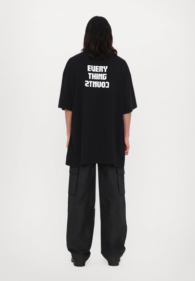 Tricou oversized negru cu text alb "EVERY THING COUNTS" pe spate, asociat cu pantaloni cargo largi negri. Material mat, croială lejeră.