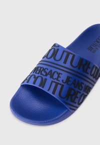 Blue rubber slide sandal featuring a wide strap with embossed black text. Text reads "VERSACE JEANS COUTURE" in a repeated pattern.
