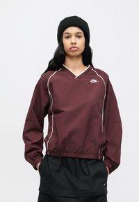 Bordový pulóverový kabátik s bielymi lemami, výstrihom do V a elastickými manžetami. Kombinovaný s čiernym dolným odevom a čiernou pletenou čiapkou.