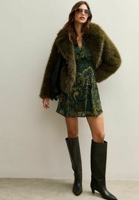 Kobieta w oversize'owej, ciemnozielonej kurtce futrzanej, wzorzystej zielonej sukience w paisley, wysokich czarnych botkach, okularach przeciwsłonecznych i z nałożonymi naszyjnikami, stoi.