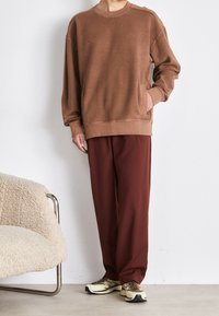 Sudadera marrón con cuello redondo y un bolsillo lateral, combinada con pantalones sueltos color marrón oscuro y zapatillas beige con acentos oscuros.