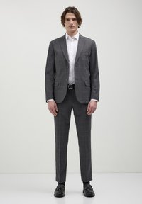 Completo grigio a motivi con vestibilità slim, caratterizzato da revers a punta, chiusura con un bottone e pantaloni coordinati. Indossato sopra una camicia bianca.