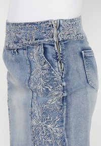Jupe en denim avec une taille haute ornée de broderies florales argentées sur le côté gauche, fermeture éclair et une poche arrière. Tissu bleu clair.