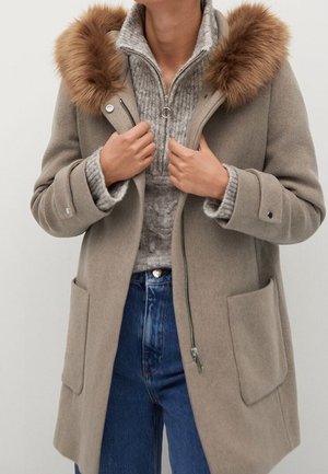 Persoon die een beige wollen jas met een capuchon met bontrand draagt, over een grijze gebreide trui en blauwe spijkerbroek, en met beide handen aan de kraag van de jas houdt.