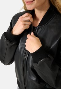Giubbotto bomber in pelle nera con polsini e collo a coste; presenta una zip frontale e una texture liscia. Inquadratura ravvicinata delle mani che chiudono la zip.